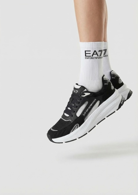 商品展示图，主体是这双黑白色的EA7 Emporio Armani运动鞋，穿在模特脚上。模特穿白色袜子，袜子上清晰印有EA7的logo。画面只展示从小腿到脚部，不出现人物身体其他部分。双脚离地有一定空预览效果
