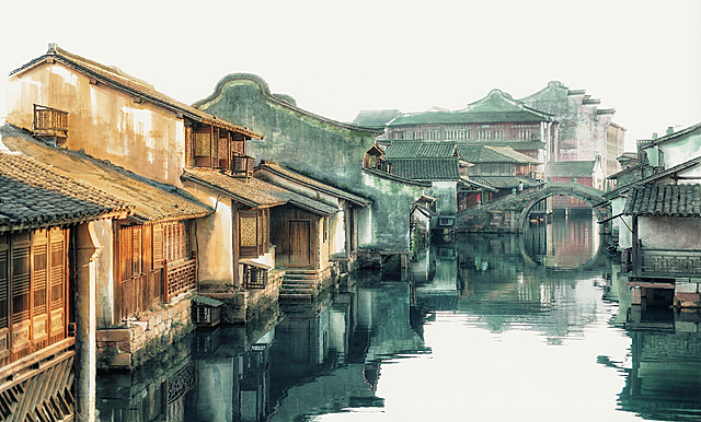 将这张江南水乡图片转换成彩色水墨风格，保留原图配色：左边房子带暖色，其他部分偏绿色，保留湖水的青色，画面保留大量留白适合做背景