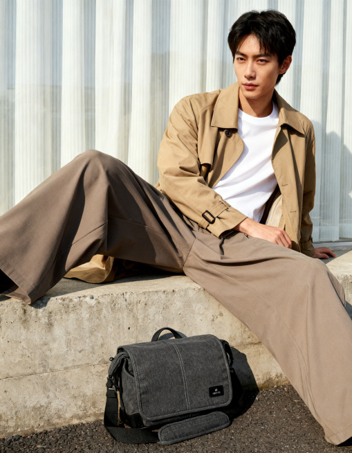 简约风户外城市商业人像摄影，电商背包产品广告场景，日韩清新画报风格，自然日常的松弛感穿搭氛围。年轻亚裔男生，卡其色风衣+白色内搭T恤+阔腿裤，韩系通勤休闲穿搭。男生斜坐水泥台阶上，主体单肩包放置在人物预览效果