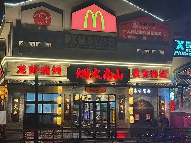 在原图中，在招牌"烟火南山"的左侧添加文字"龙虾烧烤"，右侧添加文字"夜宵排挡"，文字字体和颜色和"烟火南山"保持一致，保持原有发光效果，图片其余部分完全保留，不做任何修改，效果逼真自然
