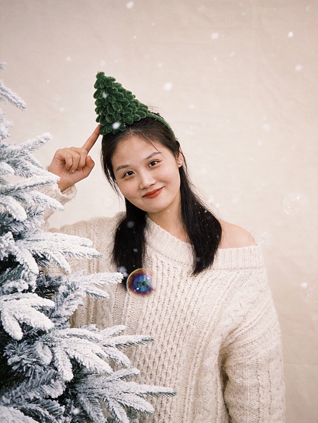 考第一张图保留人物脸型､发型､年龄特征生成圣诞主题柔和治愈系肖像写真:近景(取景范围覆盖人物头部至胸部往下5cm),居中构图,参考第二张图人物的占位和比例,人物占画面70%,平视室内场景视角(镜头高度与人物眼部平齐);50mm标准焦距,非对称均衡构图(人物偏置画面右侧黄金分割点,适度留白),人物为视觉中心;米白色粗线针织宽领落肩上衣(麦穗纹纹理,蓬松柔软质感),伪素颜清透妆(底妆轻薄保留自然肌理,浅棕眼影无眼线,自然卷翘睫毛,豆沙粉哑光唇),头部佩戴草绿色短毛绒圣诞树造型的发饰(树上点缀四颗错落分布的白色精致小圆珍珠,树顶黄色毛绒球,系白色小蝴蝶结)站姿自然随性松弛感,头微倾斜,右手举起,食指指向头顶圣诞树发饰方向,指尖舒展,浅笑注视镜头,眼神俏皮可爱,神态自然舒展,清透冷白皮甜酷精致妆容,橘红色玻璃光泽唇釉,粉桃色渐变腮红(晕染范围至眼下8mm),深棕细闪眼影+黑色上挑粗眼线+卷翘浓密睫毛,颈饰:白色丝绒质感缎带颈带蝴蝶结(带宽0.6mm,蝴蝶结尺寸2cm×2cm)漫射自然光,柔和无硬影,面部光比1:1.3,细节锐度适中(针织纹理/发丝/毛绒质感清晰可见),8K超高清分辨率,温馨感,全局饱和度+8%,叠加1mm半透明白色雪花粒子(分布密度:背景区域>人物区域),带5%胶片颗粒感(颗粒尺寸0.5px)｡暗部亮度+10%,叠加“透明彩色泡泡(直径1-3cm)”,带3%细胶片颗粒感(颗粒尺寸0.3px),浅米色哑光纯色背景布,材质无反光､表面平整,无多余纹理,前景白色喷雪仿真松枝圣诞树点缀,严格按照第一张图内容生成｡