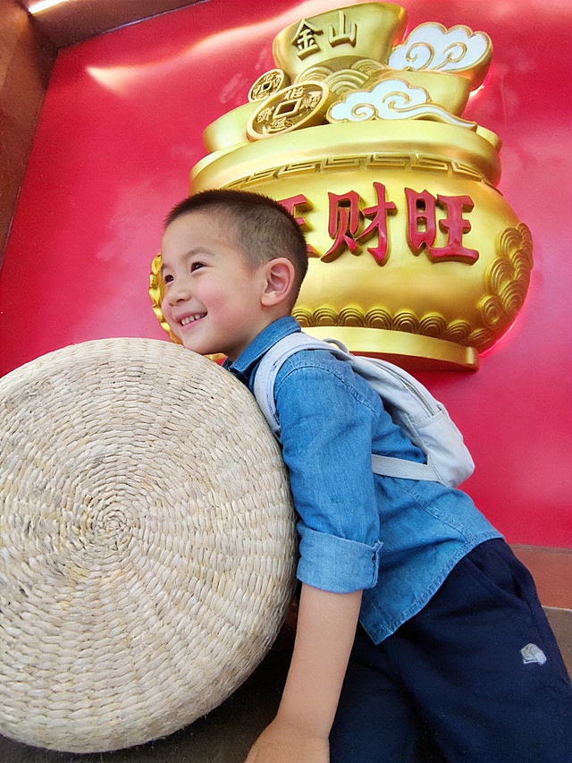 生成儿童街头打卡主题电影感摄影大片，保留参考图中小男孩的脸型、发型、年龄特征，电影叙事质感，故事感氛围，胶片颗粒质感；浓郁红金调，高饱和色彩，明暗光影对比，红墙金光氛围；真实孩童皮肤纹理，自然稚嫩肤质，孩童清爽短发；小男孩侧身趴在大草编球后，侧脸微笑露出开心表情；小男孩穿蓝色牛仔衬衫配深蓝色休闲裤，保留浅色背包配饰；室内红色背景墙，金色聚宝盆雕塑作为背景，空间简洁主体突出；中近景构图，平视视角，分辨率4k，电影级光影质感，商业人像摄影级别，画面比例3:4