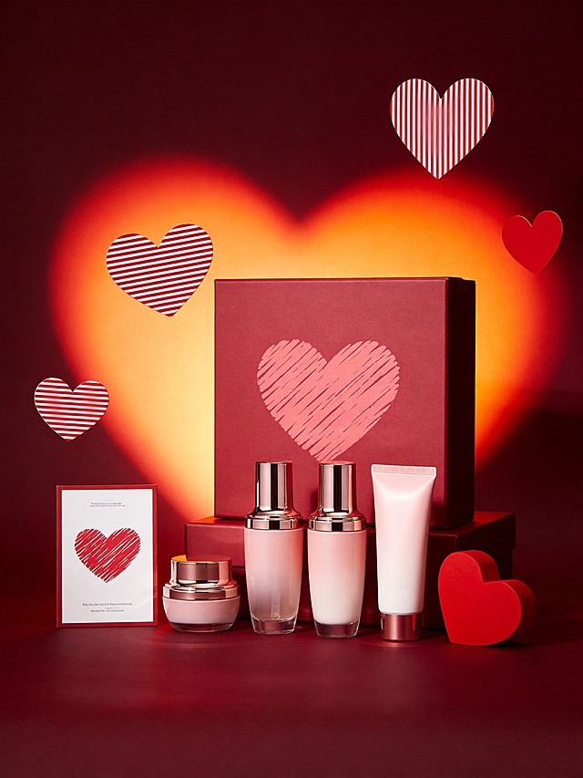 热烈浪漫风商业美妆静物摄影风格，画面以深邃酒红为背景底色，搭配渐变橙红的爱心光影作为视觉核心，整体色调浓郁热烈且富有氛围感。采用平视略俯视的视角，主体错落分布于红色平面之上：一组主体置于画面中心礼盒前偏右侧，主体后面立着同色系礼盒，礼盒上画着涂鸦爱心，呈自然直立角度，占据视觉核心区域；礼盒左侧放着展示卡，大小适中且形成稳定的视觉平衡。礼盒后面背景装饰有悬浮的红白条纹爱心与红色立体爱心道具，爱心光影采用渐变发光效果。采用侧逆光结合局部补光，营造出明暗层次丰富的热烈氛围，主体与浪漫场景融为一体，充满节日氛围感与视觉张力。