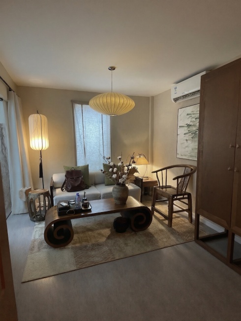 在新中式客厅的实木卷几上添加一个插有禅意干花的粗陶花瓶；在浅灰色布艺沙发上放置一个深棕色皮质背包，与整体新中式禅意暖调风格统一。保持原有场景结构和暖光氛围不变，添加的物体光影匹配环境顶光和侧光源，生成预览效果