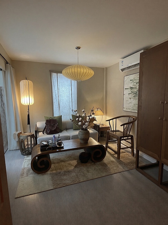 在新中式客厅的实木卷几上添加一个插有禅意干花的粗陶花瓶；在浅灰色布艺沙发上放置一个深棕色皮质背包，与整体新中式禅意暖调风格统一。保持原有场景结构和暖光氛围不变，添加的物体光影匹配环境顶光和侧光源，生成自然柔和的投影，边缘融合自然无抠图痕迹，整体风格协调。负向提示词：多余的部件、悬浮物体、比例失调、畸变透视、死黑阴影、昏暗光线、水印、文字