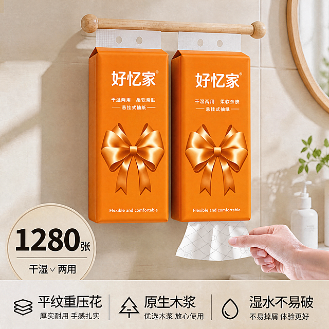 将白底图（图1）的外包装图案主题完全迁移到主图（图2）的两包悬挂式抽纸包装上：把两包抽纸正面的原有绿色卡通图案全部替换为图1的亮橙色蝴蝶结礼物主题设计，包括整个包装正面鲜艳亮橙色背景、顶部居中白色大字“好忆家”和®、下方两行白字“干湿两用 柔软亲肤”和“—— 悬挂式抽纸 ——”、下半部分中央巨大的丝绸光泽感橙色立体蝴蝶结（带高光阴影）、底部白色小字“Flexible and comfortable”；保留图2两包抽纸的包装外形、原木挂杆、挂孔、浴室瓷砖墙面、圆镜绿植洗手液场景、光线和整体尺寸完全不变，蝴蝶结光影随软包弧度调整，顶部挂孔区保留白色，文字清晰准确。