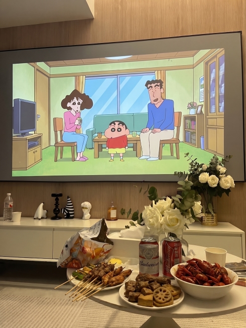 在当前客厅图片中，在圆形茶几上额外增加一些常见夜宵小吃（如烤串、卤味或小龙虾，摆盘自然），不遮挡原有的啤酒、杯子和零食包装，保持投影蜡笔小新画面和整体氛围不变预览效果