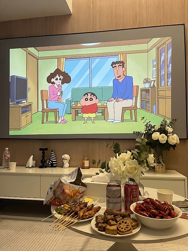 在当前客厅图片中，在圆形茶几上额外增加一些常见夜宵小吃（如烤串、卤味或小龙虾，摆盘自然），不遮挡原有的啤酒、杯子和零食包装，保持投影蜡笔小新画面和整体氛围不变