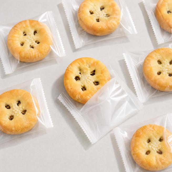 将图片中的梅干菜薄酥饼独立包装袋改为纯透明塑料材质，完全无任何文字、标签或图案；清晰展示内部酥饼实物，酥饼外观保持金黄酥脆、撒有芝麻、中心孔洞可见梅干菜馅料，与原图一致；包装袋封口为锯齿状热封边；背景预览效果