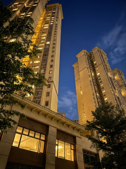 将图片中的楼盘建筑改为夜晚场景，保持原图构图和主体不变，添加高级感和质感，保留建筑轮廓和树木细节，夜晚天空深蓝色，建筑窗户透出暖黄色灯光，整体色调高级有质感预览效果