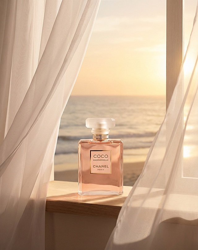 以参考图中的香奈儿COCO MADEMOISELLE香水瓶为主体，保持香水瓶的结构、Logo和文字完全不变。香水瓶放置在窗边，窗外是大海和夕阳，薄纱般的白色窗帘随风飘扬。整体为暖色调，氛围淡雅温馨，柔和的夕阳光线漫射，画面明亮通透，有前中后景层次，属于时尚画报风格的商品展示图。