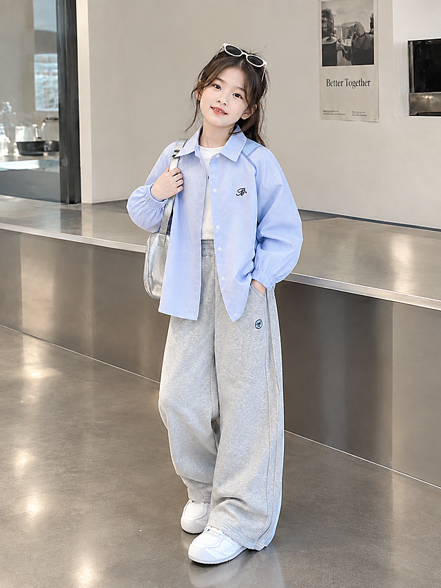 基于参考图重新生成：完整保留画面中的小女孩形象、服装造型（浅蓝色条纹衬衫、白色内搭、灰色阔腿裤、白色运动鞋、银色单肩包、头顶白色太阳镜）、姿态与微笑表情，以及现代简约室内场景和柔和自然光线；重新构图输出为3:4竖版，人物全身居中，画面上下边缘自然延展补齐，保持韩系童装摄影风格，细节清晰、画质细腻、色彩干净高级。