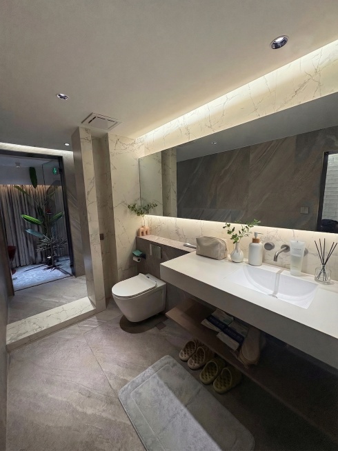 在白色洗手台的空白区域放置简约设计的洗浴用品，包括乳液瓶、洗面奶、香薰等，摆放自然有序。保持原图整体卫浴结构、光影、现有化妆包、花瓶、地毯和其他所有物品不变。光影匹配：原图光源来自顶面筒灯和镜前灯带，预览效果