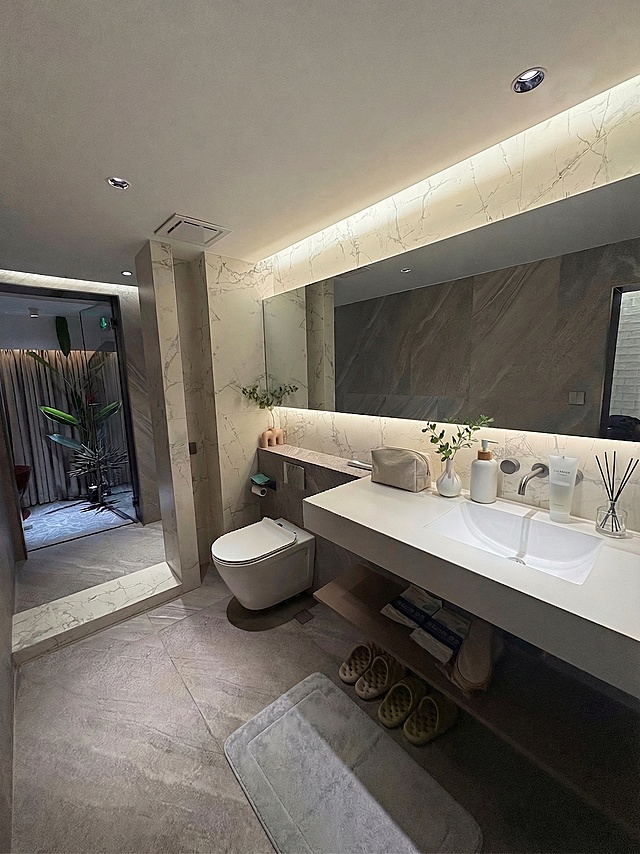 在白色洗手台的空白区域放置简约设计的洗浴用品，包括乳液瓶、洗面奶、香薰等，摆放自然有序。保持原图整体卫浴结构、光影、现有化妆包、花瓶、地毯和其他所有物品不变。光影匹配：原图光源来自顶面筒灯和镜前灯带，在新增物品对应位置生成正确的受光面、背光面以及自然阴影，阴影符合近实远虚的物理规律，整体冷调高级感统一。环境色交互：将房间的灰色大理石色调轻微映射到新增物品暗部和边缘。边缘处理：消除抠图白边黑边，使用环境色漫反射自然融合边缘。整体保持轻奢风卫浴自然真实的质感，物品放置自然协调。