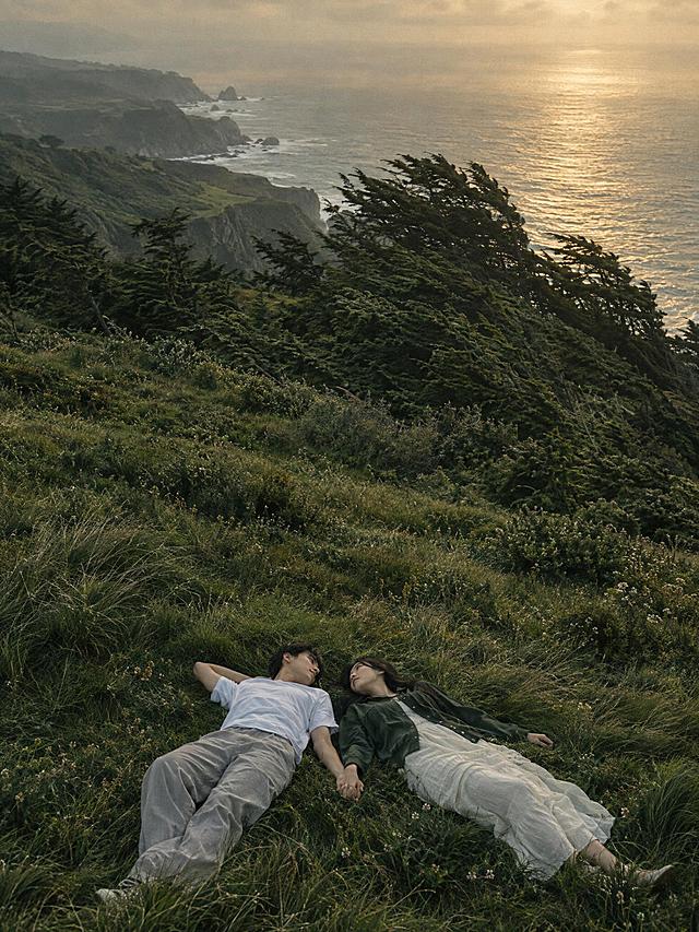 竖版3:4电影感写实静帧，复古胶片质感，低饱和墨绿色草坡与雾金色海面。宽阔的临海高地从画面下方延展到远方，画面下方一对二十多岁的年轻恋人仰躺在柔软草地上，头部相近、身体向左右展开，手在中间轻轻相握；男生穿白色短袖和浅灰长裤，女生穿深绿色薄外套和浅色长裙。中景是一片被海风吹斜的高大树丛和起伏的悬崖，左上远处隐约可见海浪与岩岸。俯视偏广角构图，安静、松弛、彼此陪伴。画面中不要任何文字、不要标题、不要字幕、不要Logo、不要水印。超高清2K画质，细节丰富。