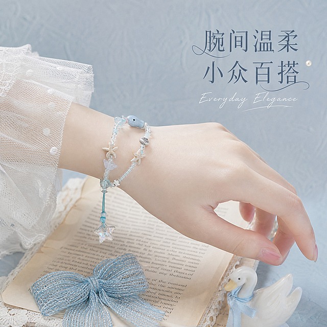 [主图-3] 佩戴效果 手模佩戴手链氛围感展示
[统一基调] 整体风格为温柔治愈芭蕾风，背景主色为浅灰蓝色带细腻肌理，标题字色为深灰蓝色，辅助字色为浅银灰色，强调色为奶油白色，字体为纤细优雅衬线体，字号层级分明，装饰元素用珍珠、丝带、蕾丝、天鹅摆件等芭蕾风元素，整体低饱和高质感，柔光影棚光。
[画面] 年轻女性手部（皮肤白皙，手指纤细，指甲为淡粉色裸色）佩戴海洋风串珠手链，手腕自然轻搭在一本打开的复古书页上，书页上放着一个淡蓝色针织蝴蝶结和一只迷你陶瓷小天鹅，背景是浅灰蓝色肌理墙面，整体为俯视45度角，手链在手腕上清晰展示佩戴效果和大小比例，手部只露出手腕和半掌，不露出面部。
[光影质感] 柔和自然光从侧上方照入，皮肤质感细腻通透，手链珠子有自然高光，金属件有柔和反光，整体光影柔和，氛围感强，无硬阴影。
[配色] 主背景浅灰蓝色，肤色白皙自然，产品浅蓝+米白+银色，文字深灰蓝+银灰，整体低饱和马卡龙色调。
[文字版式] 画面右上角放置主标题「腕间温柔 小众百搭」，纤细衬线体，深灰蓝色；下方一行小号英文手写体「Everyday Elegance」，奶油白色；文字放在空白区域，不遮挡手部和手链主体。
[输出要求] 比例1:1，文字清晰可读，产品佩戴效果自然真实，整体风格统一；禁止手部畸形、手指异常、面部出现、变形、错字、模糊、杂乱、水印、平台Logo。