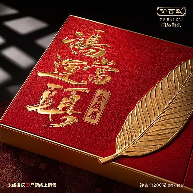图生图，基于输入的新款金骏眉红茶礼盒产品图，生成第四张主图，主题为礼盒表面烫金工艺微距特写，参考图5的风格。画面为俯视斜角微距特写，聚焦新款礼盒表面的金色“鸿运当头”烫金书法字和金色叶片浮雕，红色朱锦暗绣底纹的细腻纹理清晰可见，金色浮雕有立体凸起质感，边缘锐利高光。礼盒一角露出金色包边。下方铺暗红色团花纹锦缎。背景深黑渐变，左上角有窗格光影投射。光影为侧逆光，金色浮雕高光细腻，红色底纹肌理丰富，高级微距摄影质感。文字版式：右上角“御百载 鸿运当头”品牌标米金色。底部左右两端文字：左“未经授权 严禁线上销售”，右“净含量200克 5克×40泡”。商品主体占比约75%，画面满版无边框。1:1正方形构图，超高清商业微距摄影质感。负向提示：变形、文字错乱、产品模糊、水印、电商平台Logo。