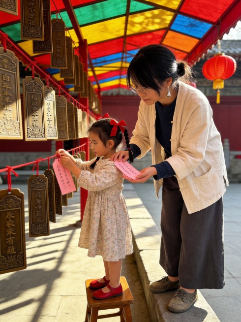 正午时分，阳光穿过南湖公园庙会的彩棚顶，斑驳地洒落在一排高悬的祈福签上。红灯笼在微风中轻轻摇曳，像一片片浮动的云霞，将整个空间染成温暖的朱砂色。阳光斜照，透过半透明的祈福卡，映出“长乐未央”“财源盈门预览效果