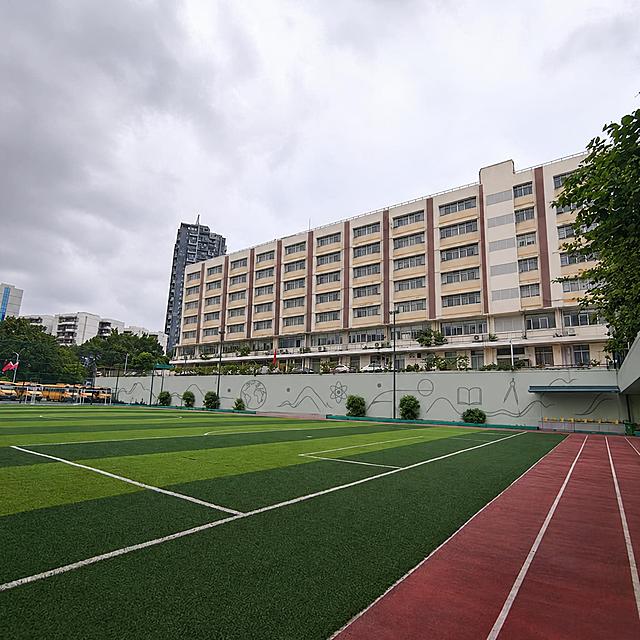 保留原照片的构图、视角、人工草坪足球场与红色塑胶跑道，只重新调整建筑与墙面配色：教学楼外立面大面积改为暖米灰（浅燕麦色），原先高饱和的明黄色带改为低饱和浅芥末黄／淡赭石的窄水平饰条，立面上的红色竖条改为灰调红棕；运动场后方原来的浅绿色挡土墙改为低饱和灰薄荷绿，去掉零散卡通涂鸦，改为极简线描的抽象地理轮廓与学科符号图案，墙面留白不少于60%，风格克制高级；天空与草坪保持自然真实。画面中不出现任何文字、字母、数字、标语、校名、logo。