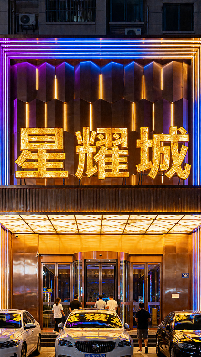 夜晚城市里的商务KTV门头，招牌是巨大的金色发光字体 “星耀城”，门头装饰有暖黄色 + 蓝紫色渐变霓虹灯光，建筑外立面是金属质感的装饰面板，门口停着几辆轿车，有几位顾客进出，整体氛围奢华亮眼，金属质感墙面搭配玻璃门，夜晚灯光效果华丽，画面色彩明亮、对比度高，质感出众，高清细节，8K