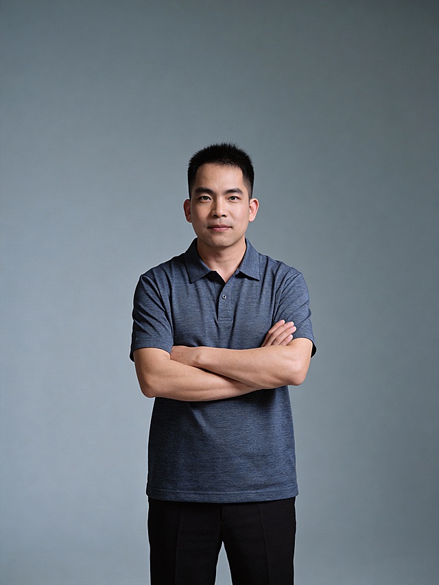 参考第一张图保留人物脸型､年龄等特征生成职场形象照,参考第二张图人物占位和比例,标准焦段50mm,F/4光圈(F/2.8-F/4区间),平视视角(镜头与人物视线齐平);人物占画面70%,人物居中(居中构图),位于画面中心,为视觉聚焦点,入境范围头部及以下至臀部下方10cm(半身像);左右留少量背景空间;头部与画面顶部保留5%空间;头部发型完整;头肩比例协调,身穿偏蓝的深灰色精纺面料polo衫短袖(翻领样式),上衣自然垂落于裤装之外,呈现出随性且规整的垂坠状态;黑色西裤,款式简约大方,日常商务休闲的风格,双臂交叉于胸前姿态,肢体自然松弛;呈正面抱胸的自然站立姿态:左臂搭于右臂上方,肩颈自然松弛下沉,姿态自信从容｡正式严谨专业稳重,身形匀称,姿态端正｡发梢微微修整干练大气,主体锐利,影棚侧光布光(主光来自画面前方),明暗过渡柔和,背景光线均匀,强化色彩对比的高级感｡简约偏冷调灰色纯色背景(呈现均匀柔和质感),极简干净;32K画质超高分辨率､细节清晰,整体风格雅致､兼具现代精致感;肤质精细化处理;眼神直视镜头,目光坚定且澄澈,表情自然.嘴角上扬;面部光比1:1.5,4800K中性光,微微修饰脸型,肤质哑光细腻,面部雾面柔光,轻透裸肌感,真实毛孔纹理,32K画质,去除人物之外的所有元素｡