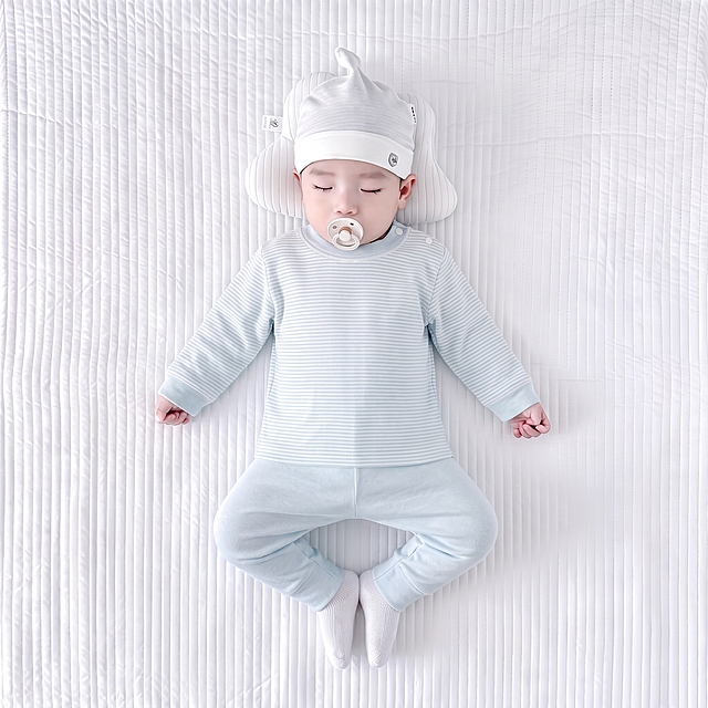 婴儿服装产品图，婴儿仰卧在白色绗缝床垫上，穿着浅蓝色细条纹长袖上衣和纯色浅蓝长裤。上衣胸口没有徽章，条纹连续平整。裤子面料光滑无纹理。整体画面温馨自然，商业摄影风格，高清细节。