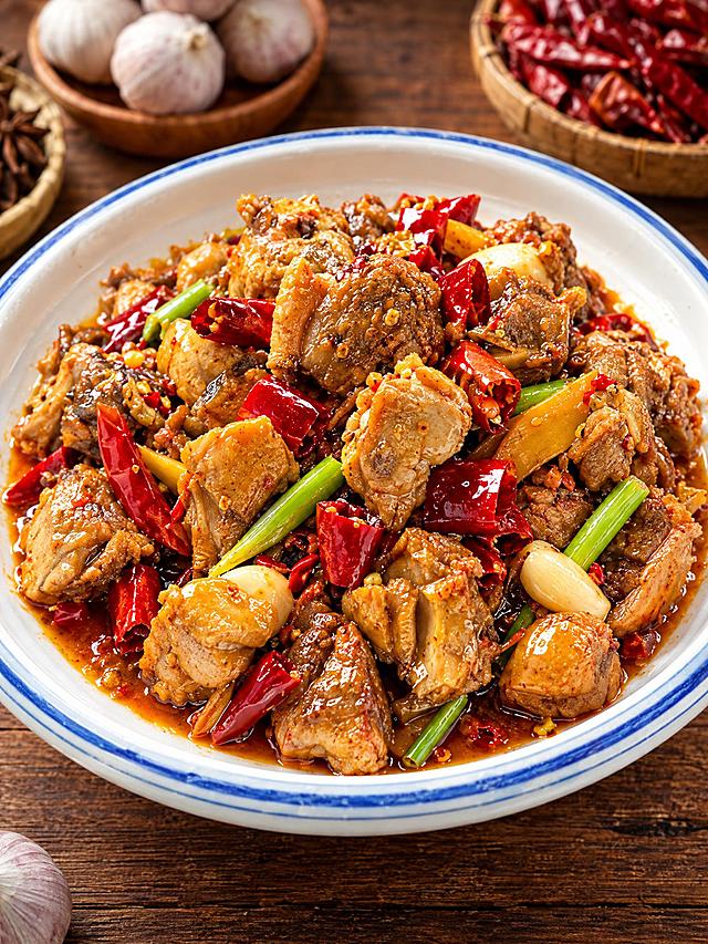 基于参考图中的红烧鸡块菜品，生成30度俯拍特写角度的美食摄影图，聚焦菜品细节，保持菜品色泽红亮、辣椒葱段蒜瓣配料丰富，蓝边白瓷盘装盛，木质桌面背景，暖色调专业美食摄影，高清细节，诱人食欲