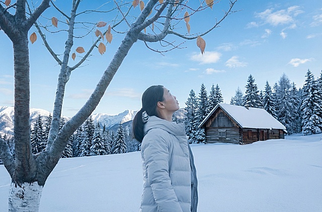 背景中改成积雪覆盖的地面、树木、一座小木屋以及远处的山脉，天空呈蓝色并飘着少量云朵，整体色调以蓝白为主，营造出清冷纯净的冬日氛围。人物服装的蓝色与白色雪地形成和谐的色彩搭配