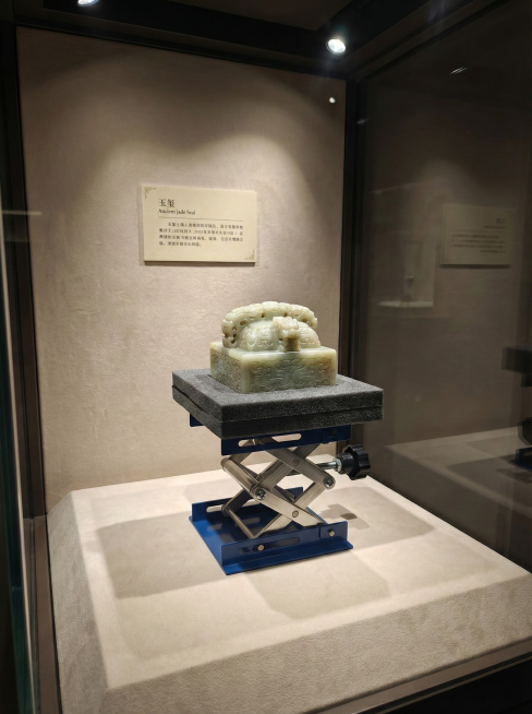 博物馆展柜内，小型升降台托着一枚古代玉玺，玉玺放在定制海绵衬垫上，上方有聚焦射灯，背景是浅色绒布和说明标签，玻璃反光处理，庄严静谧，高清文物摄影 比例3:4预览效果