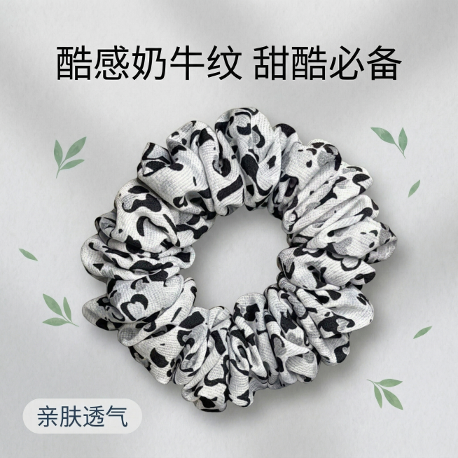 基于图一的奶牛纹大肠发圈，保留产品外观与原图一致。参考图二的创作风格：浅灰色纯色高质感背景，发圈居中摆放，主体占比适中，搭配简约绿色叶片装饰在角落，不遮挡产品。文字排版参照图二：上方大标题 "酷感奶牛预览效果