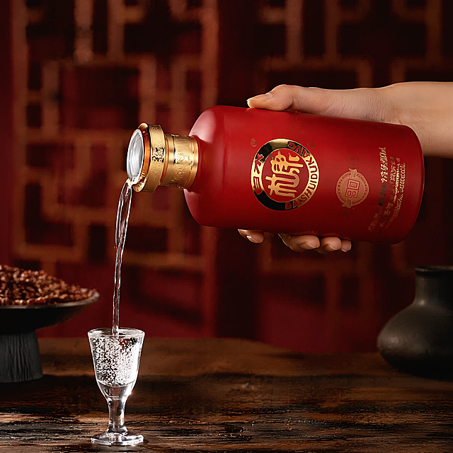 白水杜康窖90红色瓶白酒，严格100%还原上传商品图的产品外形、包装、LOGO、印刷文字、净含量，完整保留原图中的瓶型、轮廓、结构、比例、瓶盖、标签版式、品牌logo、口味文字、容量信息、材质质感、红色瓶身颜色与正面朝向，包装、配色、产品结构、瓶身造型与标签布局均与上传图保持一致，产品不变形，文字无错漏，画面不添加任何文字，不添加酒盒。
参考图为白酒倒酒商业拍摄，复刻保留新中式轻奢动态场景，中国女性手部握持对应红色瓶身酒瓶，向下方玻璃小酒杯稳定倒酒，动态自然不冻帧，产品和动作匹配准确，无任何文字。
色彩体系：红金暖棕调，红色瓶身在暗红色背景衬托下更突出，酒体通透视觉。
场景光影：中国女性手部握持酒瓶，向下方玻璃小酒杯倒酒，白酒液体自然流动动态，深棕色木质台面，背景模糊红色中式屏风，侧逆光突出酒体质感，酒水通透有光泽，自然立体光影，产品主体准确对应红色瓶款。
严格锁定宽高比为 1:1。
4K 分辨率，写实商业摄影，版式精美，产品主体清晰，画面无任何文字。