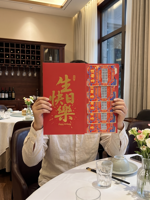 严格1:1还原原构图与红包样式，人物姿势和坐姿朝向不变：人物面部完全被左右并排展开的超大生日红包遮挡，无露脸；红包为左右两页并排展开的样式，左页是烫金繁体生日快乐文字，右页是五张喜相逢刮刮乐彩票，完全预览效果