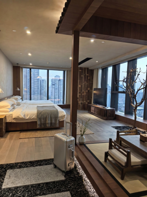 在房间地面上放置一个银色带轮子行李箱，在木质柱子旁边放置一个小花瓶，在床铺上放置一个毯子预览效果