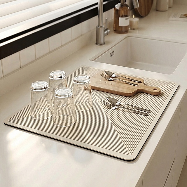 现代美式厨房浅色石英石台面场景，背景虚化保留水槽、龙头、少量餐具，主推图案沥水垫（16"×20"）平铺于台面靠近水槽处，上面放置刚洗好的玻璃杯、刀叉、一个小砧板，呈现正在使用的状态。自然晨光从左侧窗照入，产品表面有柔和高光，体现硅藻泥细腻颗粒感，杯底无水渍，垫子边缘干净利落与台面无缝贴合，整体色调温暖整洁有序，符合minimalist but functional审美