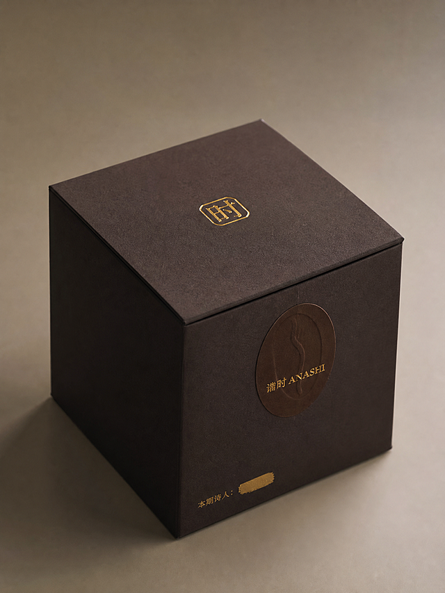 方形翻盖礼盒，尺寸12cm×12cm×12cm，材质为深檀色特种纸，表面哑光磨砂质感。盒盖正中央有一枚微凸烫金工艺的金色"时"字印章LOGO，盒身贴椭圆形咖啡豆轮廓封口贴纸，哑光特种纸材质，印着烫古铜金文字"谙时 ANASHI"，礼盒四面侧面留白，其中一个侧面印有极小文字"本期诗人：———"，名字被可刮开金色涂层覆盖，整体展示角度为斜45度，质感高级简约，光影柔和突出材质