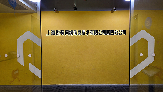 将图片中黄色墙面上的“万物新生”立体发光字替换为“上海悦契网络信息技术有限公司第四分公司”，保持原有的黑色立体字样式、背后发光效果、字体风格、厚度和光影完全一致，文字大小与入户门墙面比例协调自然，符合公司入户门名称展示的视觉效果，图片中其他所有元素（玻璃门、墙面纹理、监控摄像头、地面、灯光等）保持不变，整体画面真实自然，无修改痕迹