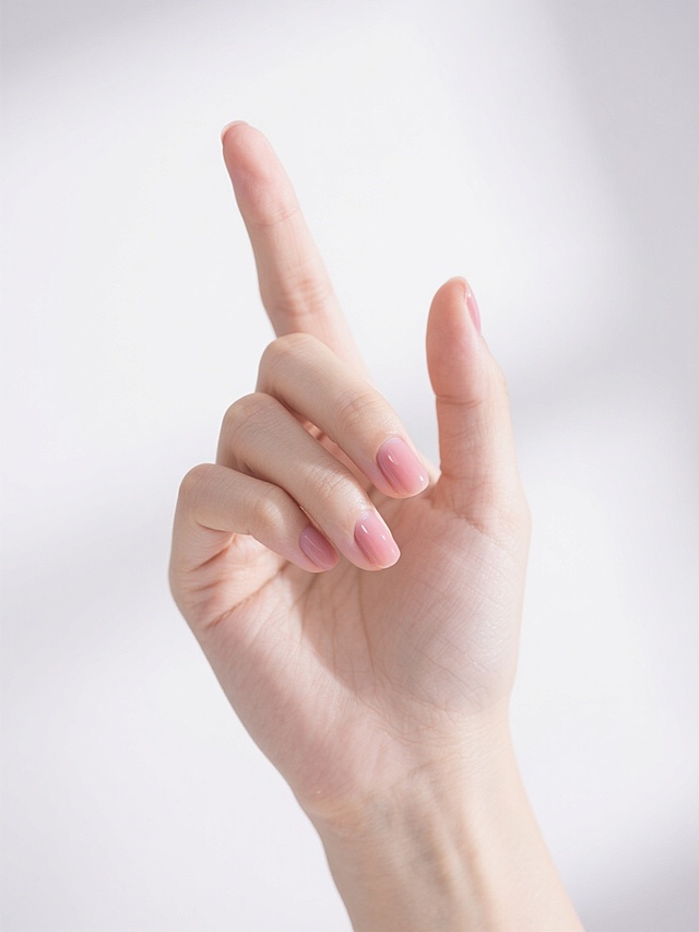 一只年轻女性的手，手指修长，指尖涂有淡粉色指甲油，手部皮肤细腻光滑，呈现自然的肤色，手背有轻微的血管纹理，手呈食指向上伸直、其余手指自然弯曲的姿势，手腕部分露出，背景为纯白色，光线柔和均匀，阴影浅淡，整体画面简洁干净，具有产品展示级的高清质感和写实风格。