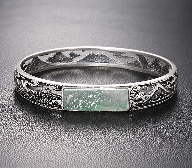 Macro jewelry photography, soft diffused studio lighting, subtle shadows, ultra realistic texture, high resolution, luxury product shot, neutral background. Generate a closed round sterling silver bangle featuring an arched mountain carving gemstone (25mm length, 8.2mm width) set horizontally at the center. The gemstone resembles a sculpted mountain ridge with elegant flowing curves. The bangle is made of 925 sterling silver with intricate openwork (filigree cut-out) patterns inspired by traditional Chinese landscape painting. Flowing water motifs and layered mountain silhouettes wrap around the entire bracelet, forming a continuous scenic composition. The silver surface has partial oxidized antique finishing to enhance depth and contrast, creating an ink-wash painting effect. The mountain carving is half-bezel set with an open gallery underneath for light penetration. Style: Oriental artistic jewelry, poetic, refined, cultural elegance, high-detail craftsmanship.