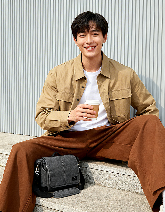 年轻亚洲男性，面带微笑，身穿卡其色外套、白色内搭和棕色阔腿裤，手持咖啡杯，坐在户外石阶上，身旁放置深灰色帆布MATIN品牌主体单肩包，现代简约户外场景，浅灰色竖条纹背景墙，清新自然的时尚生活摄影风格，明亮柔和的日间自然光，均匀通透的光线，中景构图，色彩柔和温暖，日系简约风格，高清晰度，质感细腻，轻松惬意的都市生活氛围