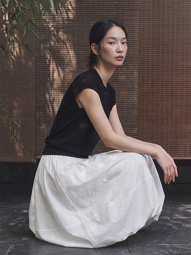 【主体保护指令】请保持参考图1中的【同一模特、同一服装、同一场景光感】一致，包括面部五官、服装细节、背景色调、光影，不做改动。仅姿势、镜头景别、朝向按描述重新生成。

画面整体为电商模特展示图，模特蹲姿，双手自然搭在膝头，身体微微前倾，目光望向镜头，表情清冷，中景景别，3/4侧朝向，高角度机位，自然构图。

模特的面部五官、发型、肤色、气质、身形与参考图1保持一致，不做改动。

服装以参考图1为准，完整保留款式、颜色、材质、剪裁、图案、版型、细节等一切细节。上衣为黑色圆领短袖针织衫，领口处有一枚细小的弧形金属饰件，袖口为罗纹收边，针织纹理细腻柔软，下摆罗纹收边；下装为白色大摆茧型半裙，高腰松紧腰头，裙长及小腿中部，裙身蓬量饱满呈茧型弧度，裙子中下部分有一条横向缝线分割，裙摆弧度宽大圆润，自然垂落。

背景与参考图1保持一致，为棕色竹帘背景搭配竹叶光影，色调统一。

光影与参考图1保持一致，柔和自然的侧光，竹叶光影斑驳，明暗过渡细腻。

画质与参考图1保持一致，超写实细腻质感。

整体风格为东方极简高级时装摄影风，清冷疏离的高级感，低饱和色调，电影质感，杂志大片调性。

中景景别，50mm标准焦段，高角度俯拍机位，人物居中，画面留白自然。

画面与参考图1在模特面部、服装、背景、光影、色调上保持高度一致；人物特征稳定（五官/肤色/发型一致）；服装细节准确；关节合理、姿势自然。