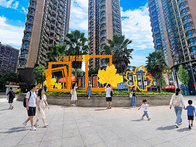 在保持原图所有建筑、橙色画框装置、枫叶造型、绿化、广场地面、天空及其他所有元素完全不变的前提下，在广场地面区域增加一些散步的亚洲市民，包含成年人和儿童，人物自然分布，姿态放松；同时在绿化中的橙色画框和枫叶造型装置旁增加一些正在打卡拍照的亚洲游客，人物与场景融合自然，光影透视与原图一致，整体画面真实自然，不改变原图其他任何内容和构图