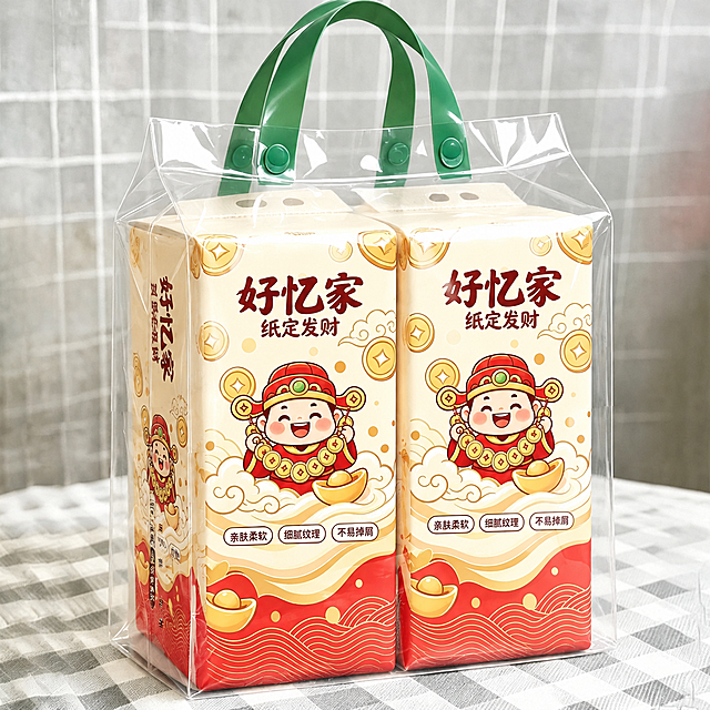 将第一张白底图中的好忆家财神包装图案完整迁移到本图中两提悬挂式卫生纸的包装正面和侧面上，保留本图的透明手提袋、绿色提手、格子桌布背景等所有环境元素不变。包装上完整保留：好忆家品牌名、"纸定发财"字样、卡通财神形象、金币元宝祥云元素、红白金配色、底部波浪纹、"亲肤柔软/细腻纹理/不易掉屑"三个标签。包装形状、透视角度、光影与原图一致，图案贴合包装曲面自然不变形，透明袋的反光和折射效果保留。