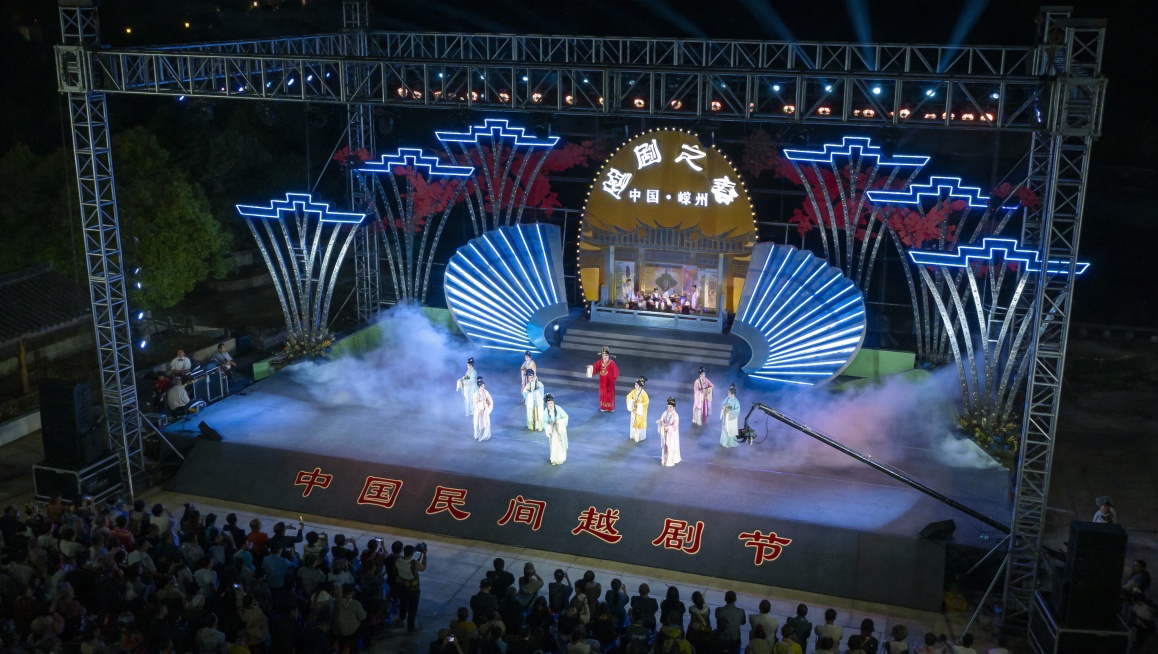 参考图中的舞台结构，画出一个露天夜间舞台，鸟瞰，舞台上能看见有一些人在表演越剧，舞台背景是大大的字是“越剧之春”，下方标注地点是“中国 嵊州”。舞台下方还有大大的字“中国民间越剧节”，很多观众预览效果