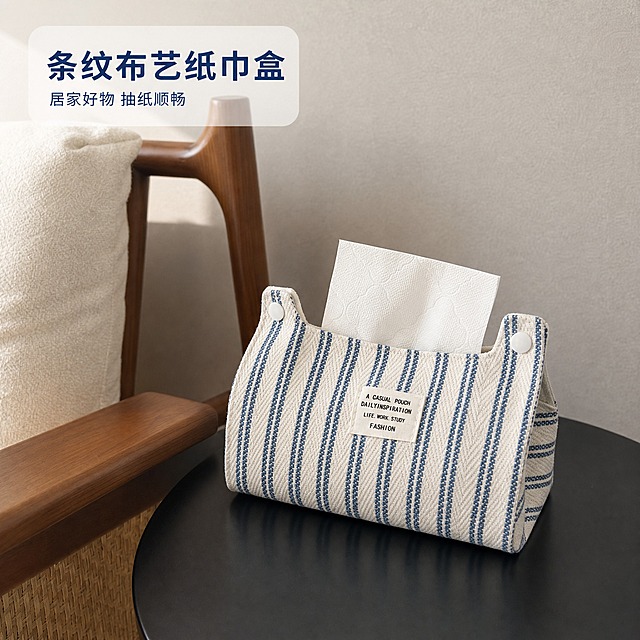 [主图-1] 蓝白条纹布艺纸巾盒 空间氛围单品呈现主图
[统一基调] 温馨居家生活感 自然柔和午后光线 米灰暖色调 高级材质透视 画面干净有呼吸感
[画面] 产品为蓝白竖条纹帆布纸巾盒，正面有白色布标写着"A CASUAL POUCH DAILYINSPIRATION LIFE. WORK. STUDY FASHION"，两侧白色按扣设计，盒内抽出一张白色压花抽纸，纸巾样式与图二保持完全一致。产品放置在黑色圆形小桌面上，画面左侧保留图一背景中的实木椅子扶手与米白色布艺靠垫，后方为浅灰色肌理墙面。产品居中偏右摆放，主体占画面约65%，45°平视微俯视角，完整展示产品正面与侧面轮廓。椅子与靠垫作为环境陪衬，不抢主体视觉。
[光影质感] 商业摄影质感 午后自然光从左上方照入 产品左侧受光略亮 右侧形成柔和自然阴影 桌面有轻微接触倒影 帆布材质纹理清晰可见 布标有轻微立体感
[配色] 主色米灰+蓝白条纹 背景浅灰墙面 木色椅子 黑色桌面 整体低饱和暖调 蓝白条纹为视觉焦点
[文字版式] 左上角放置轻量标题文字"条纹布艺纸巾盒" 字体为简洁无衬线粗体 深蓝色字 下方小字"居家好物 抽纸顺畅" 白色半透明底托 文字不遮挡产品主体 占比小于20%
[输出要求] 1:1正方形 2K分辨率 产品清晰不变形 纸巾样式与图二完全一致 无多余杂物 无水印 无平台Logo 无错字