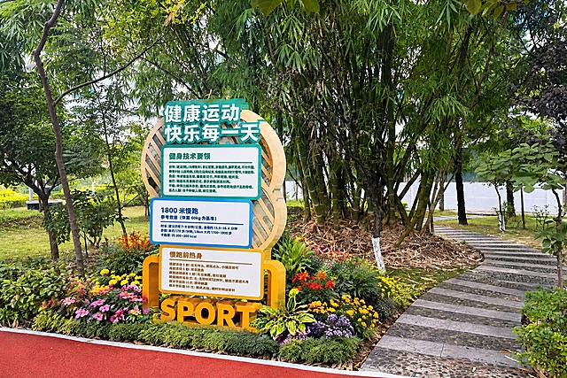在雕塑下方栽种小型花卉，雕塑底部周围种植各色小型观赏花卉，包括粉色、黄色、紫色、红色等多色小花，搭配低矮绿色观叶植物，形成精致的花境效果，花卉自然环绕雕塑底座。其余场景保持不变，竹林背景、石板步道、红色塑胶跑道，白天自然光，真实光影效果，3:2横构图